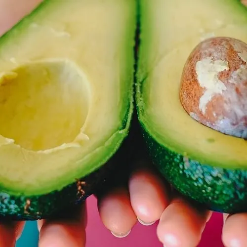 Manos sostienen dos mitades de aguacate cortadas, una con hueso, sobre fondo rosado vibrante