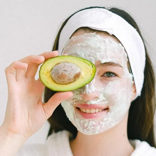 Mujer con mascarilla facial blanca sostiene medio aguacate frente al rostro y sonríe