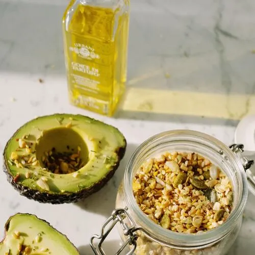 Mitad de aguacate junto a una botella de aceite y un frasco con semillas sobre mesa de mármol