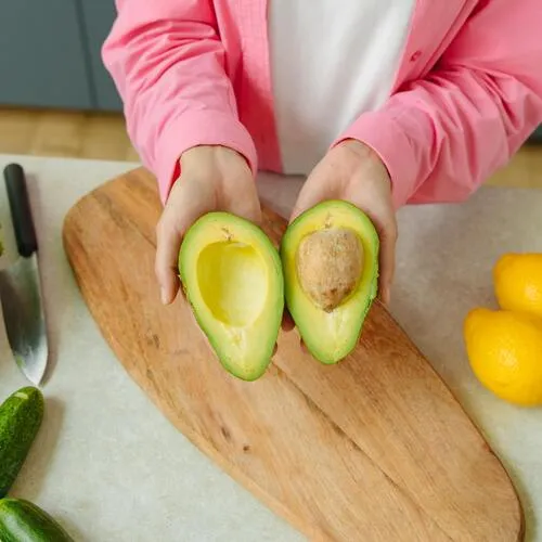 Mujer con camisa rosa sostiene dos mitades de aguacate sobre una tabla de cortar de madera