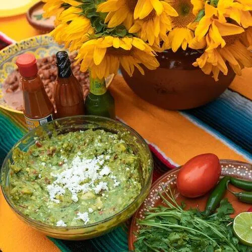 Bowl de guacamole con queso fresco encima, salsas picantes y un jarrón con girasoles sobre mantel mexicano