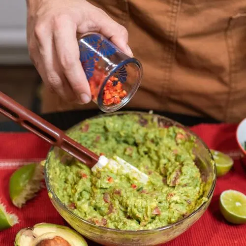 Mano agrega ají rojo picado a un bowl grande de guacamole sobre mantel rojo, con limones al lado