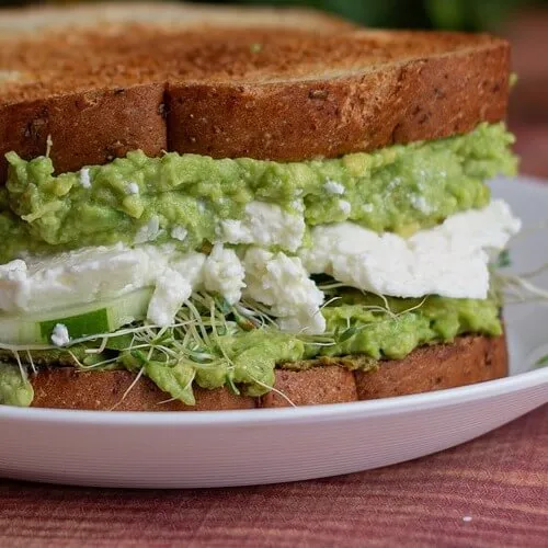 Sándwich de pan integral relleno con guacamole, queso feta, pepino y brotes
