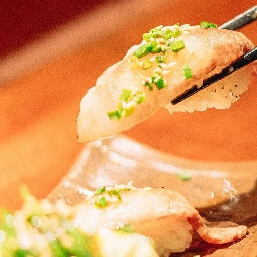 Pieza de sushi sostenida con palillos, decorada con cebollín y semillas, fondo cálido